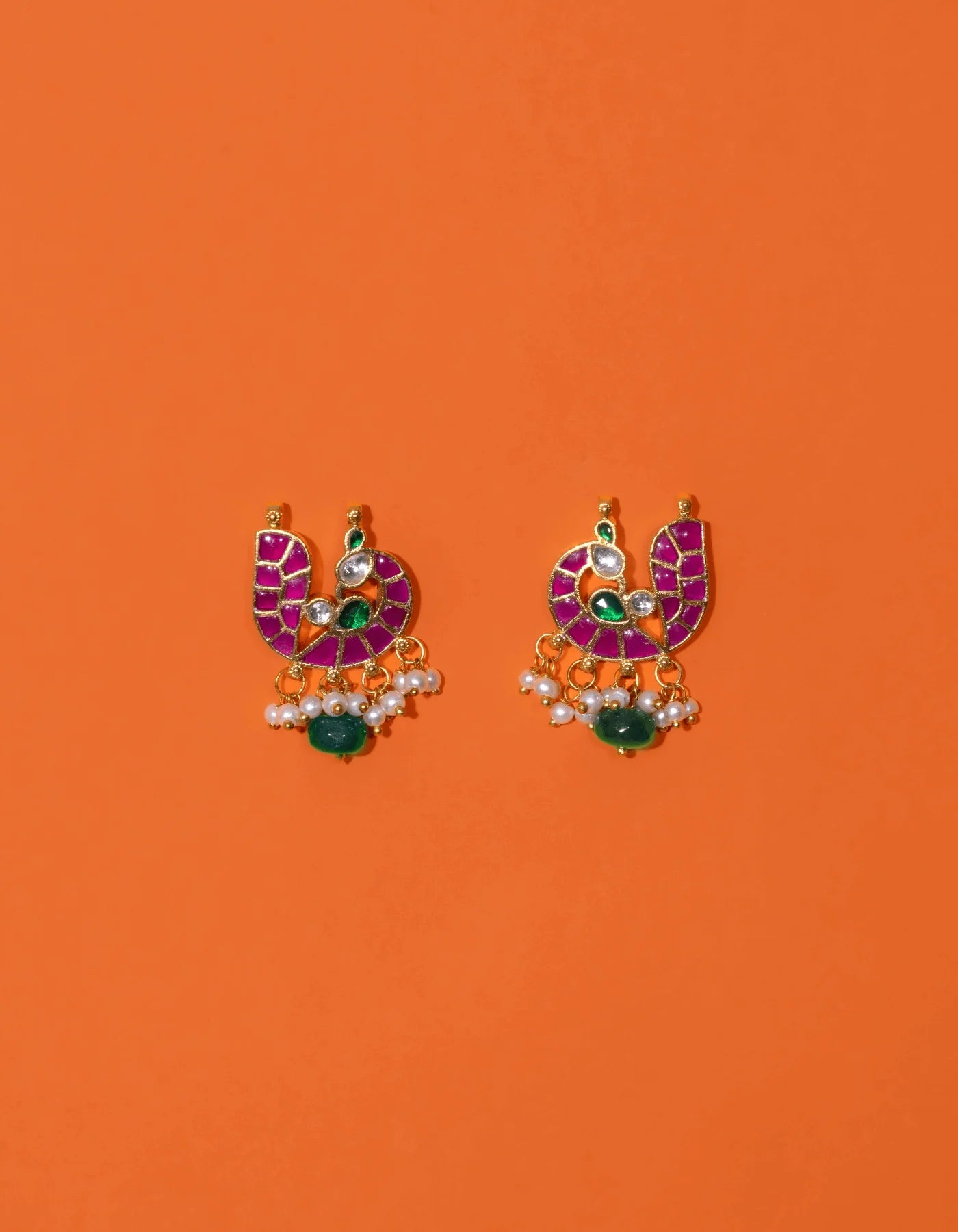 Artistic composition of hands holding decorative gold earrings adorned with radiant pink panels, green gemstone touches, and layered pearl embellishments.