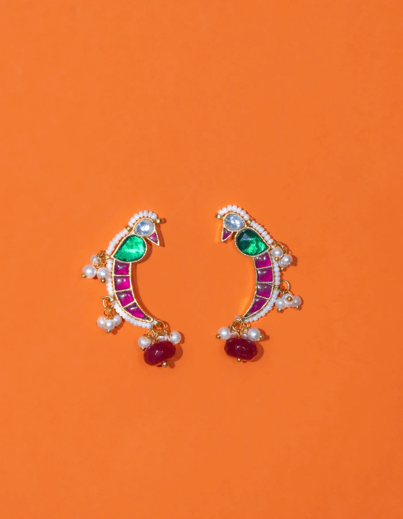 Artistic close-up of handcrafted earrings with magenta panels, emerald-toned stones, and delicate pearl fringes resting on a pigment-coated hand.