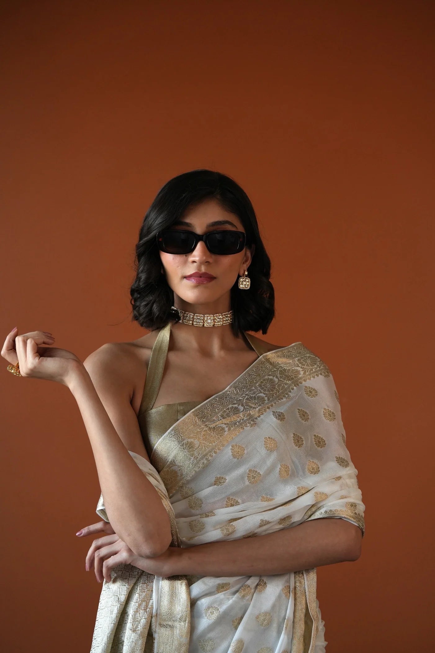 Portrait of a woman wearing a gold saree with intricate patterns, styled with a diamond choker, statement earrings, and black sunglasses against a warm-toned background.