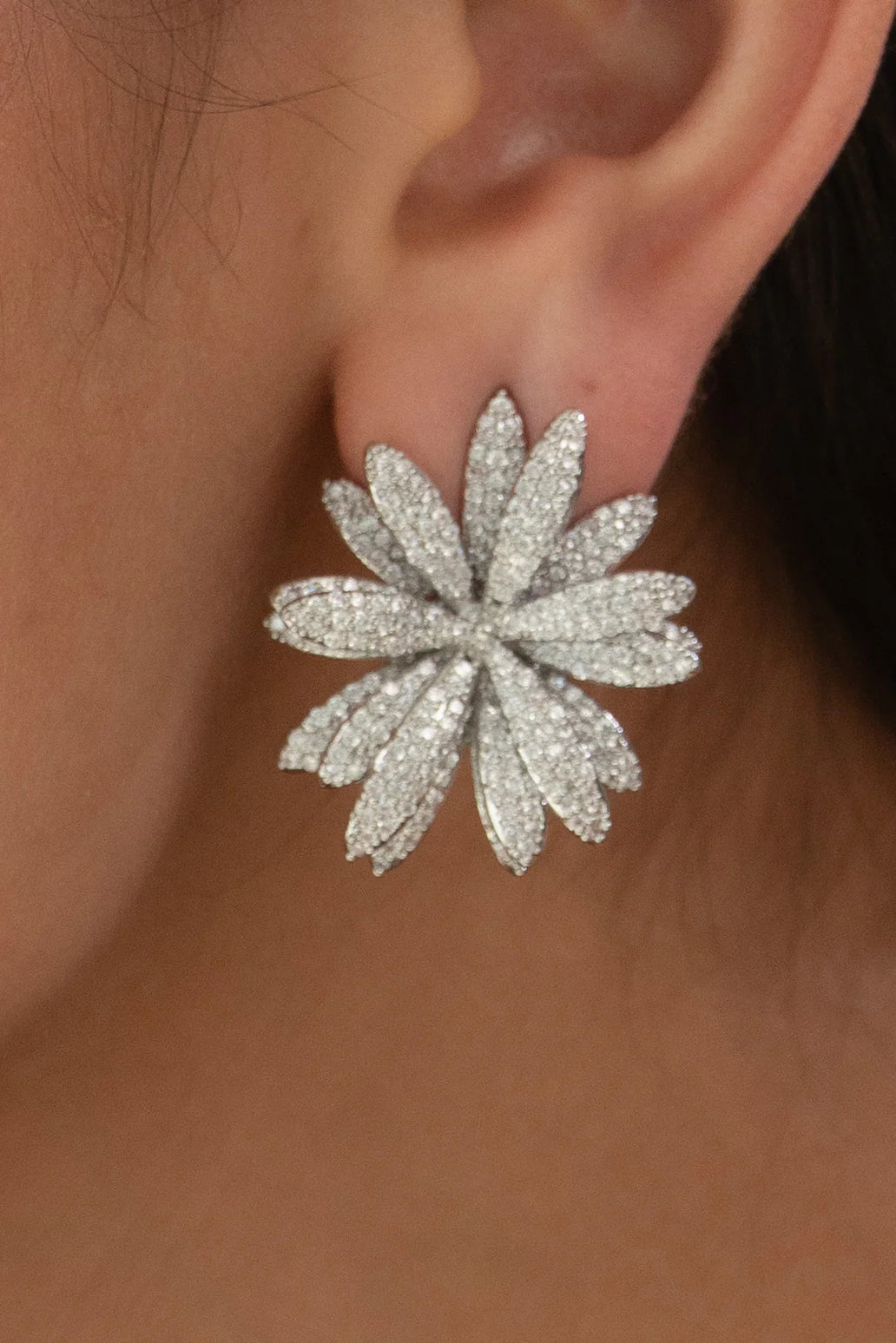 Close-up of a person’s ear wearing a large, flower-shaped stud earring encrusted with small sparkling stones.
