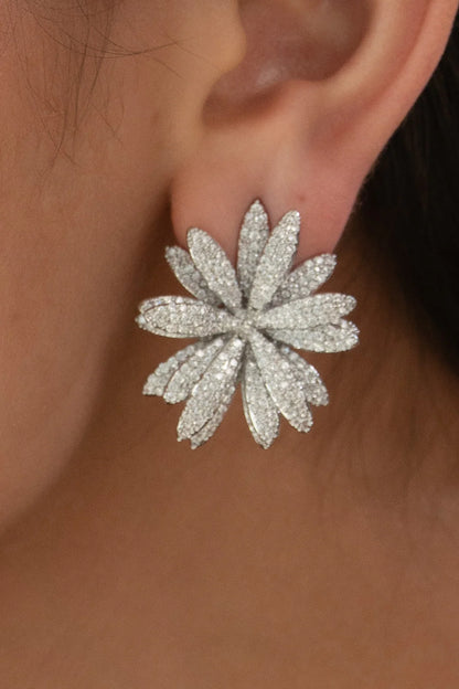 Close-up of a person’s ear wearing a large, flower-shaped stud earring encrusted with small sparkling stones.
