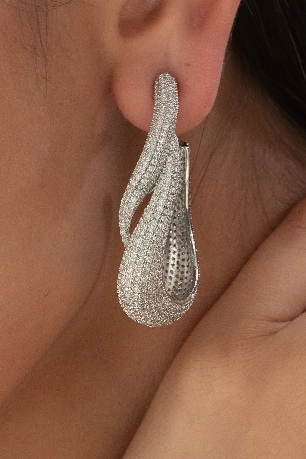 Close-up of a person’s ear adorned with a sculptural silver drop earring featuring a flowing, looped design and densely set sparkling stones.
