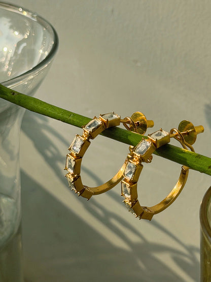 Elegant gold hoop earrings with rectangular crystal stones displayed on wooden rod in soft sunlight
