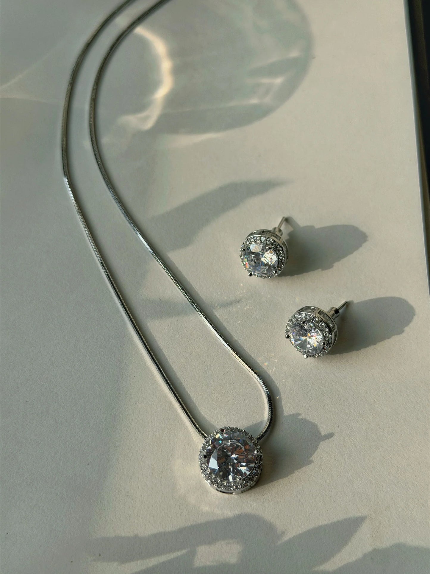 Elegant silver necklace with round crystal pendant and matching stud earrings on soft light background
