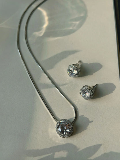 Elegant silver necklace with round crystal pendant and matching stud earrings on soft light background
