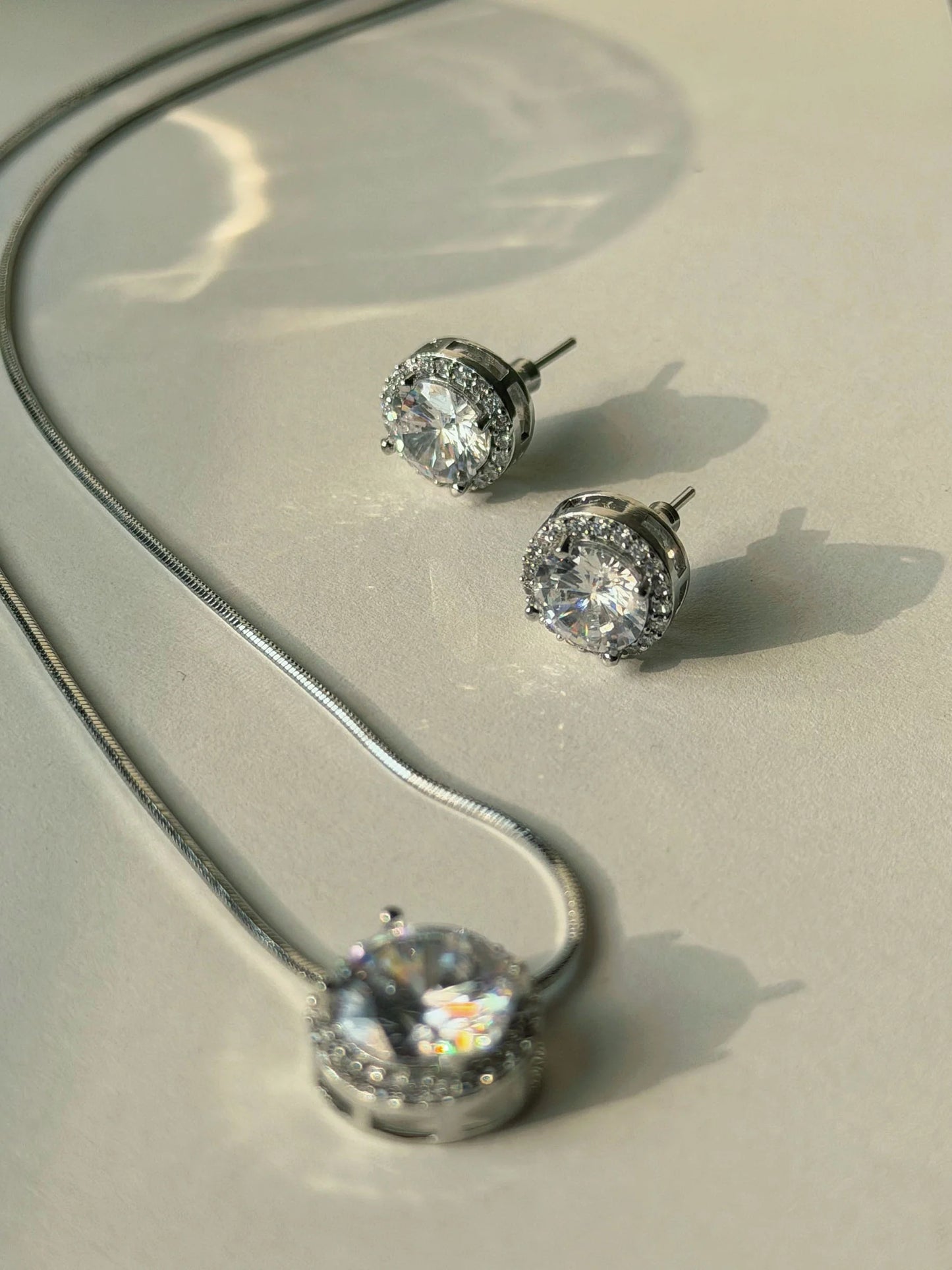 Classic silver jewelry set featuring round diamond-like pendant and stud earrings on neutral surface
