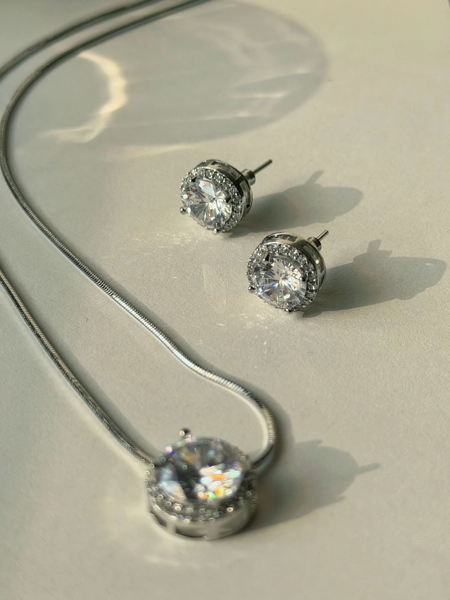 Classic silver jewelry set featuring round diamond-like pendant and stud earrings on neutral surface
