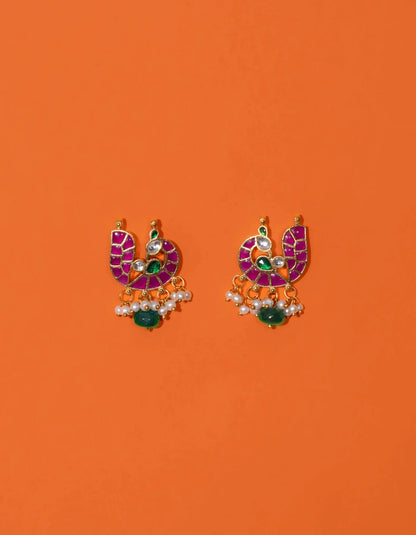 Artistic composition of hands holding decorative gold earrings adorned with radiant pink panels, green gemstone touches, and layered pearl embellishments.
