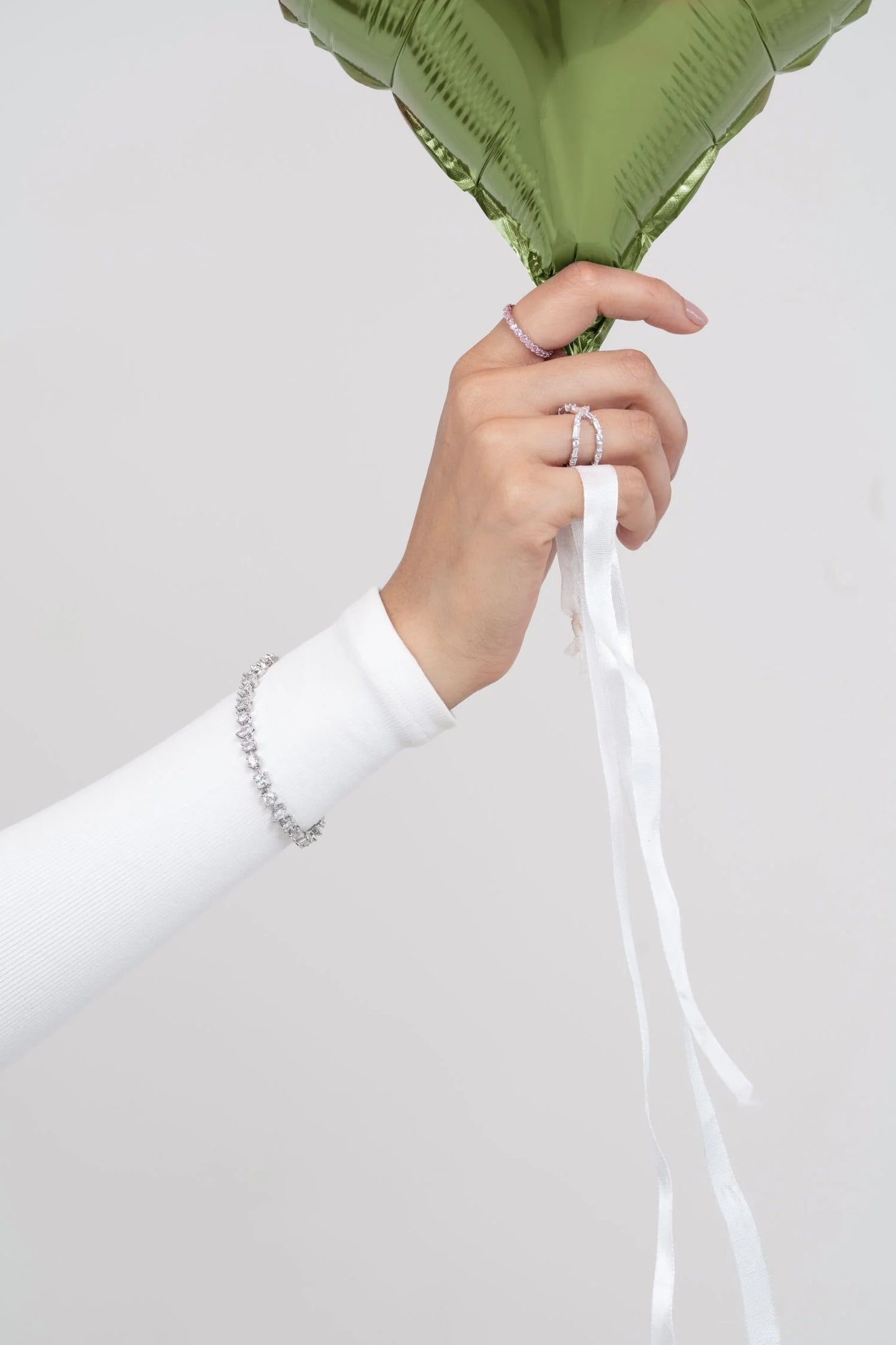 Close-up of a hand holding a green balloon ribbon, wearing delicate diamond rings and a slim diamond bracelet on a white sleeve.
