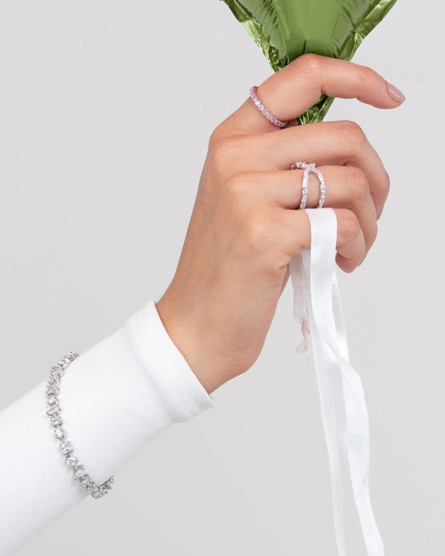 Minimal styling shot of a hand with fine diamond jewelry holding a glossy green balloon against a clean background.
