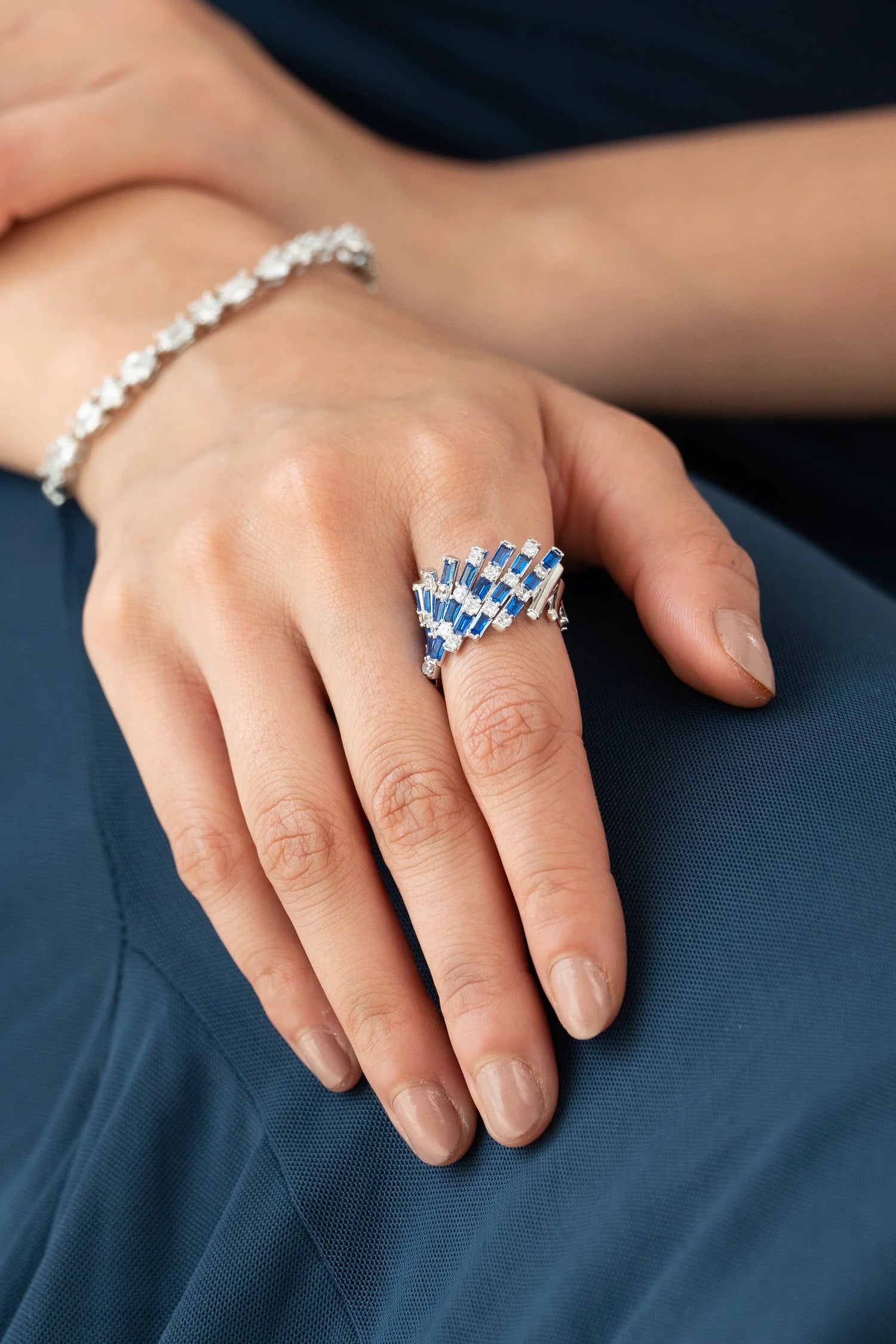 Close-up of a hand wearing a statement sapphire and diamond ring with cascading baguette-cut blue stones, paired with a delicate diamond bracelet.

