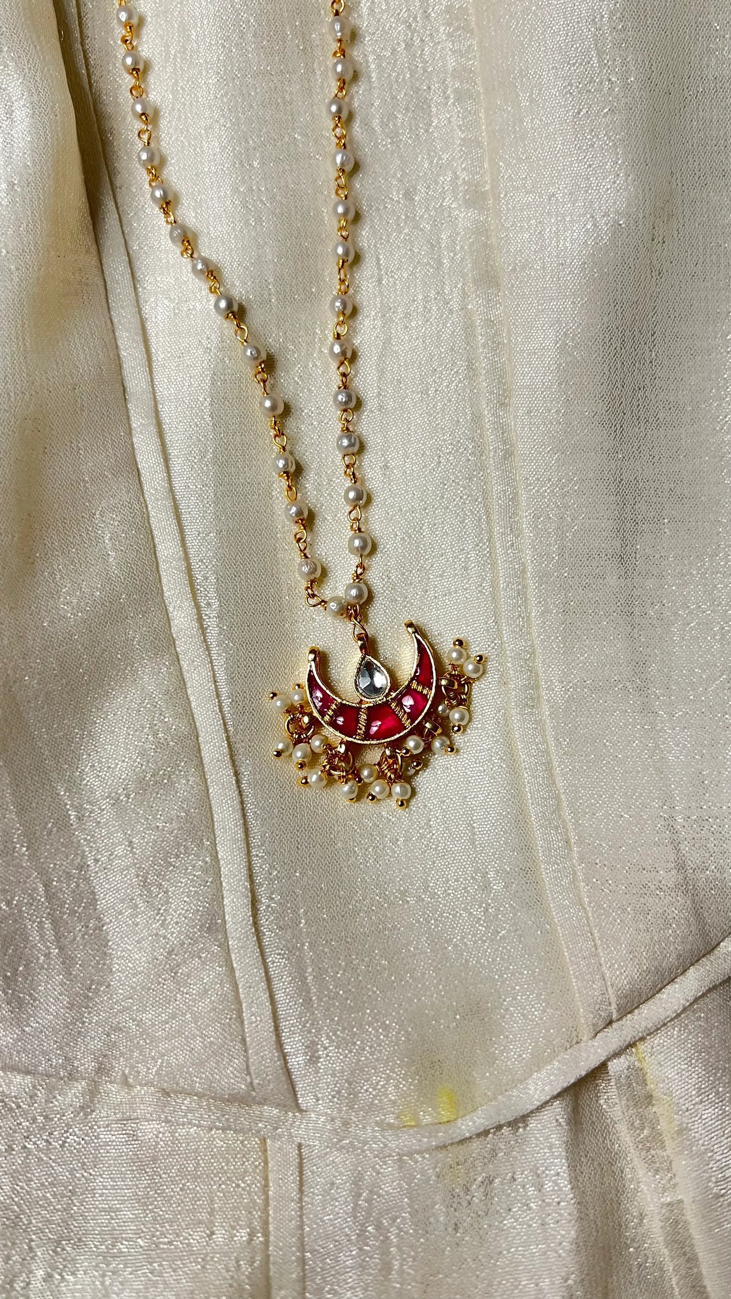 Refined jewelry detail of a gold and pearl chain paired with a statement red pendant featuring delicate hanging pearls.

