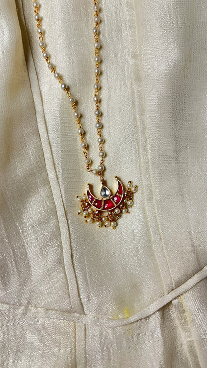 Refined jewelry detail of a gold and pearl chain paired with a statement red pendant featuring delicate hanging pearls.

