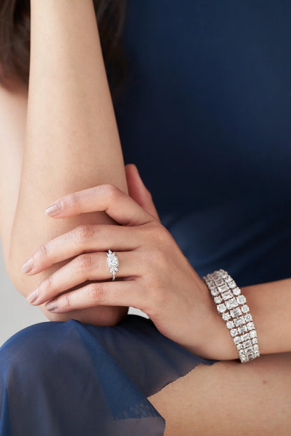 Close-up of hand and wrist adorned with a sparkling diamond ring and a wide geometric diamond bracelet against soft blue fabric.
