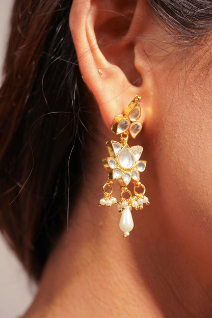 Close-up of a woman wearing a delicate gold necklace with small dangling charms and matching floral drop earrings, styled with a deep blue outfit.
