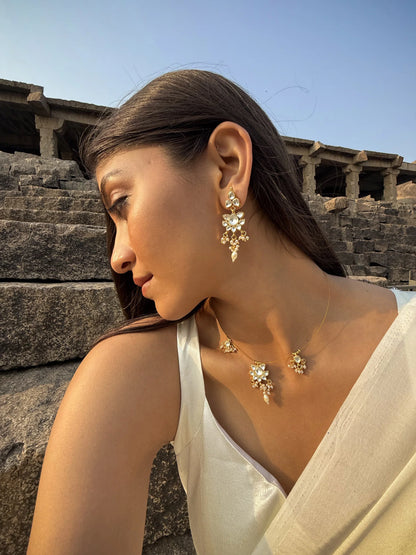 Refined close-up of a woman smiling softly while wearing a delicate charm necklace and matching floral-inspired earrings.

