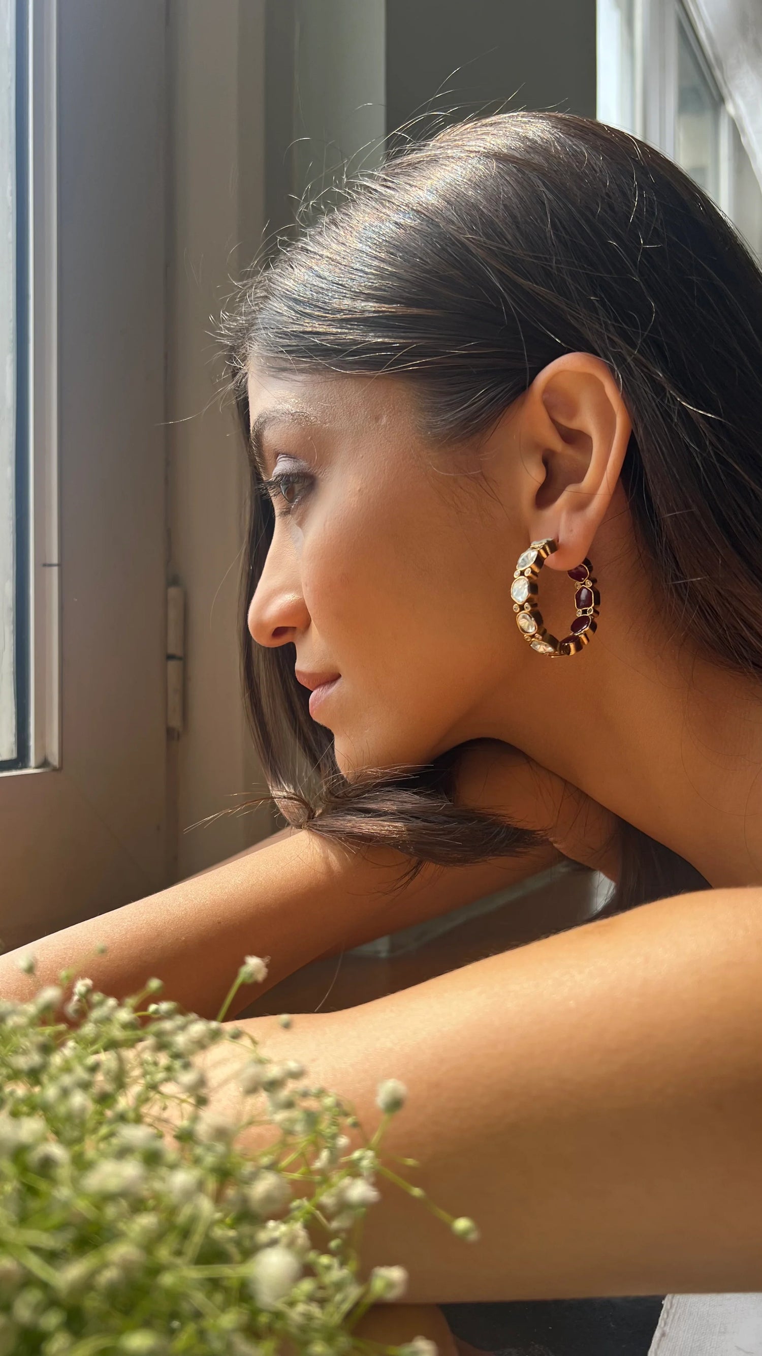 Fine jewelry close-up showcasing a circular earring with mixed gemstone accents worn by a woman near a window.
