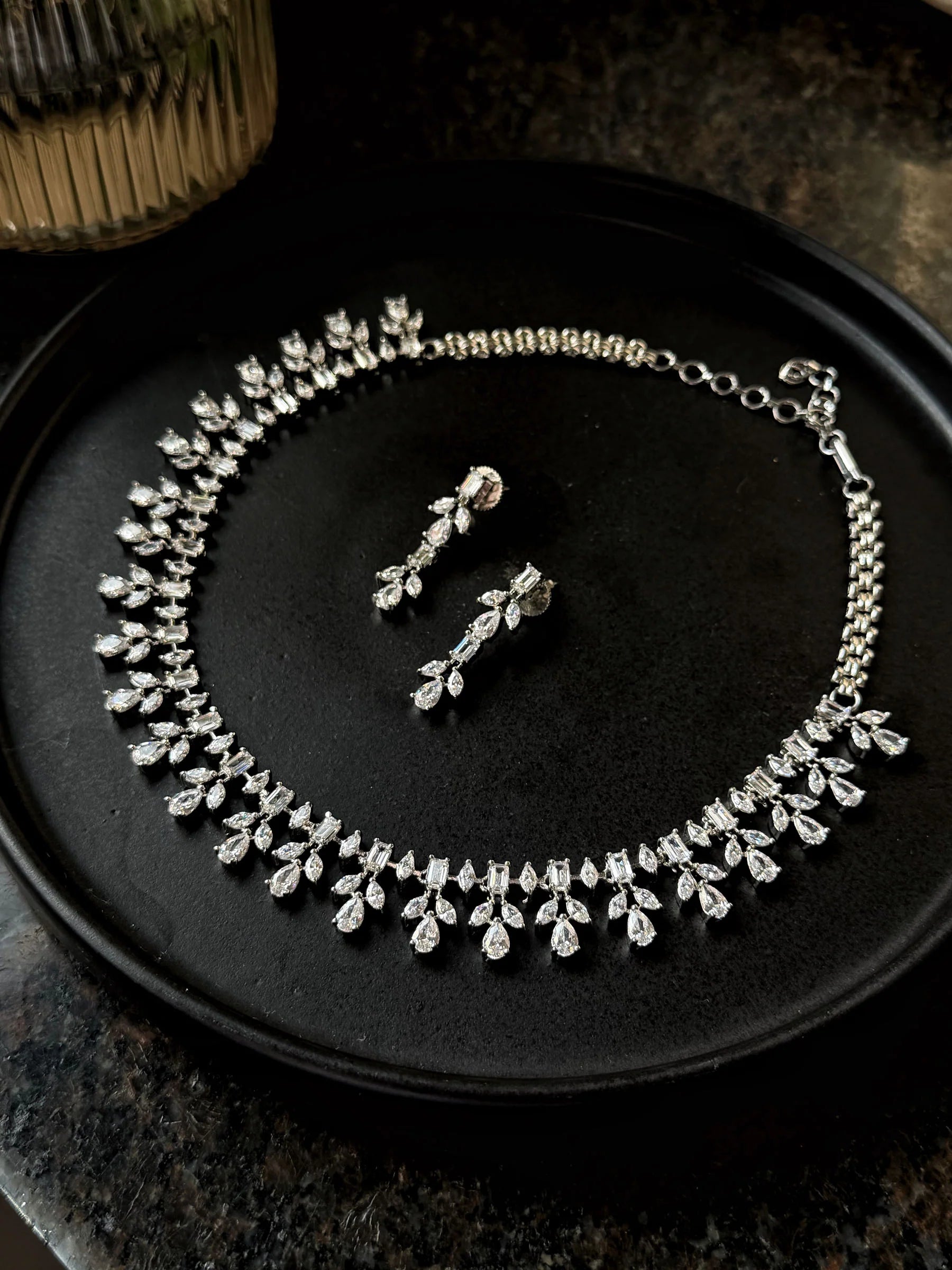 Elegant silver necklace and drop earrings set with sparkling crystal stones on black background
