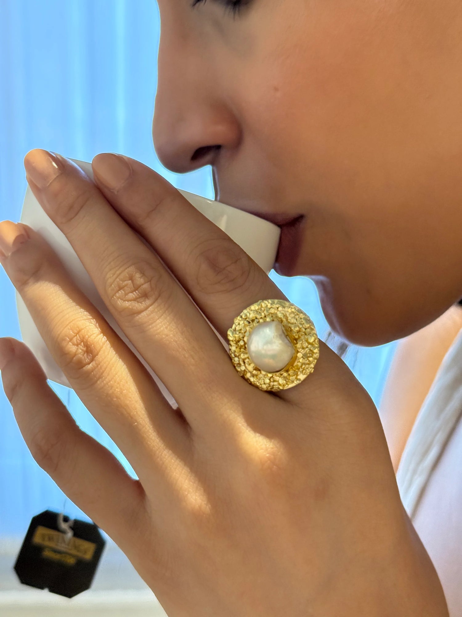 Luxury pearl statement ring on hand as woman drinks from white cup in cozy setting
