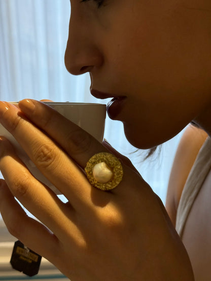 Woman enjoying coffee wearing oversized gold ring with pearl detail in warm lighting
