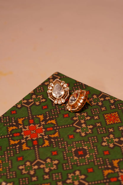Elegant gold stud earrings with teardrop crystal stones and halo design on patterned fabric background

