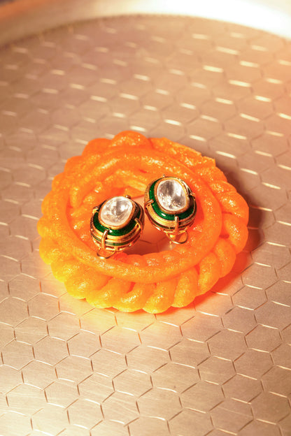 Close-up of hands holding bright orange jalebi with a pair of gold and green oval earrings featuring clear stones placed in the center.
