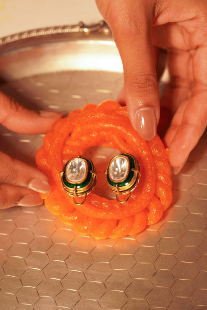Close-up of gold statement earrings featuring oval clear stones with green enamel detailing, styled on a vivid textured orange surface for a bold, vibrant contrast.
