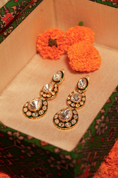 Hands presenting a vibrant jalebi with decorative gold and green earrings featuring oval crystal-like stones placed in the middle.
