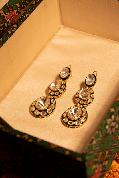 Close-up of glossy orange jalebi held between fingers, with a pair of traditional gold earrings featuring green enamel and oval clear stones displayed at the center.
