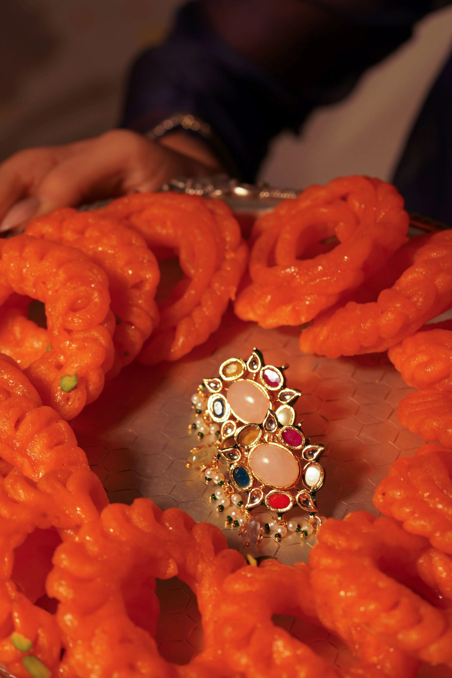 Traditional jalebi dessert used as a display base for elegant gold earrings with green enamel and oval transparent gemstones.
