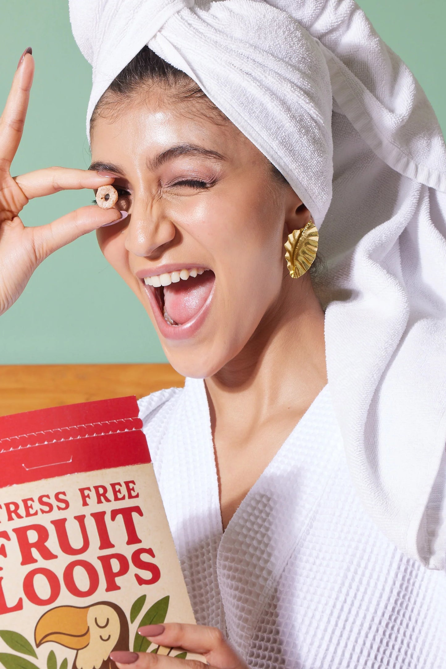Playful woman in a white bathrobe and towel holding cereal near her eye, wearing bold gold statement earrings
