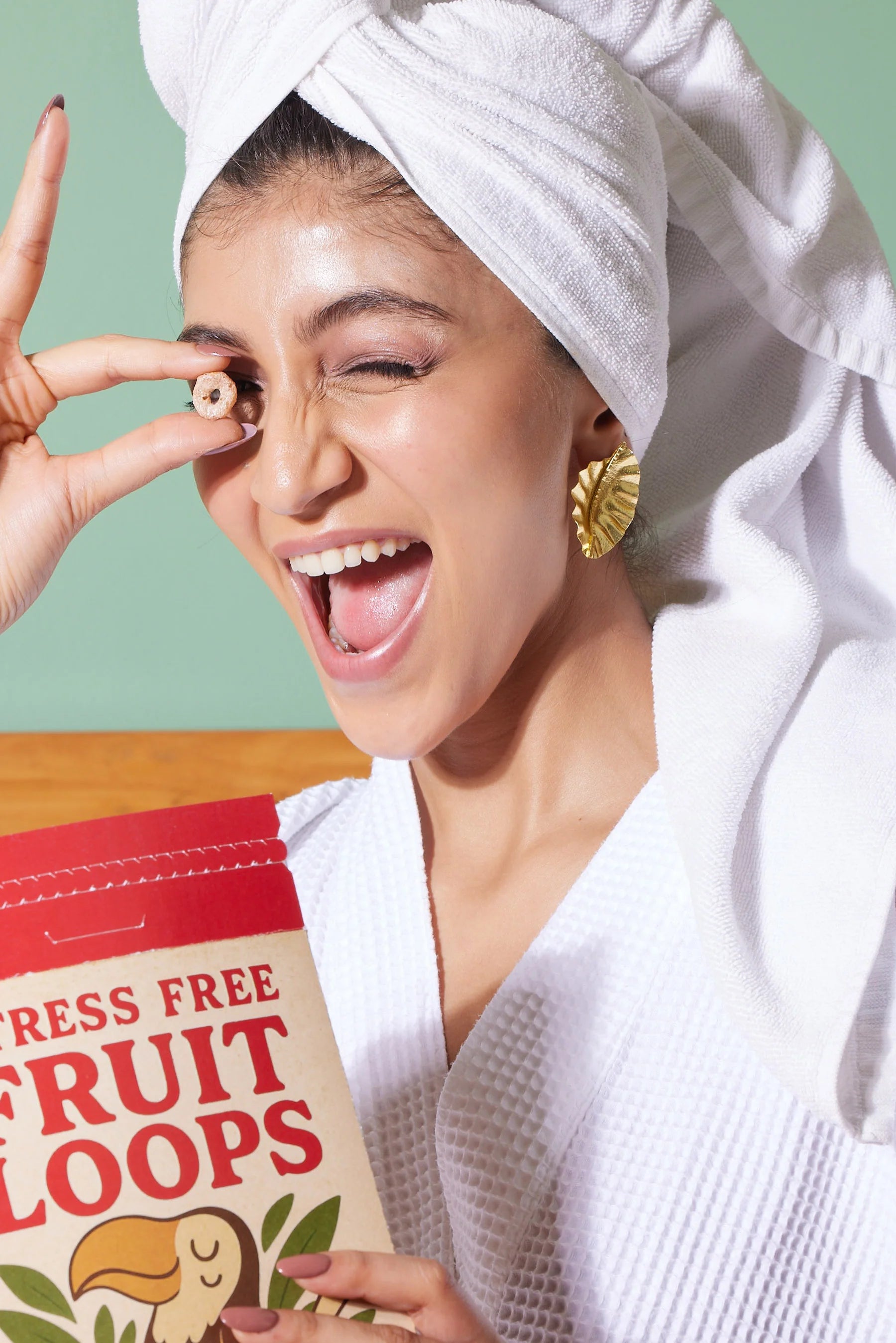 Playful woman in a white bathrobe and towel holding cereal near her eye, wearing bold gold statement earrings
