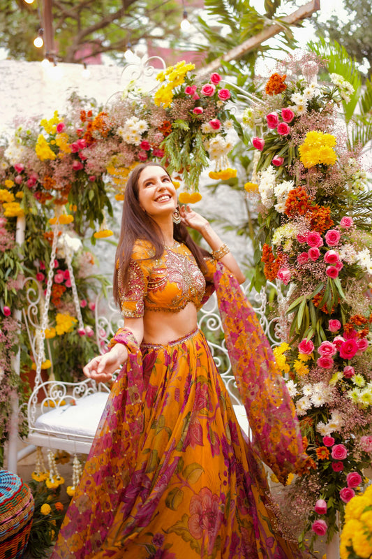 Woman in vibrant yellow floral lehenga posing under a colorful flower arch at a festive outdoor setting
