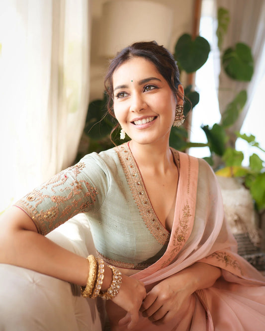 Smiling woman in pastel green blouse and peach saree wearing traditional gold earrings and bangles indoors
