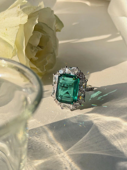 Luxury emerald ring with rectangular green stone and surrounding diamonds on neutral surface

