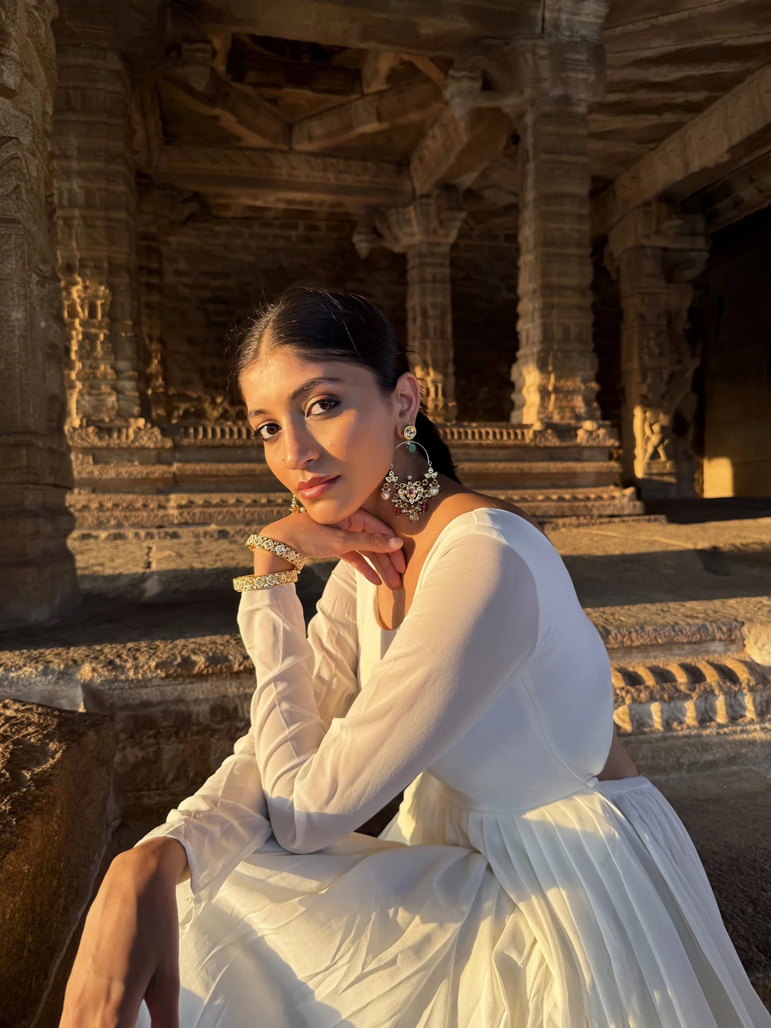 Close-up of a woman in a flowing white outfit wearing ornate chandelier earrings and bangles, seated against intricately carved stone temple architecture at golden hour
