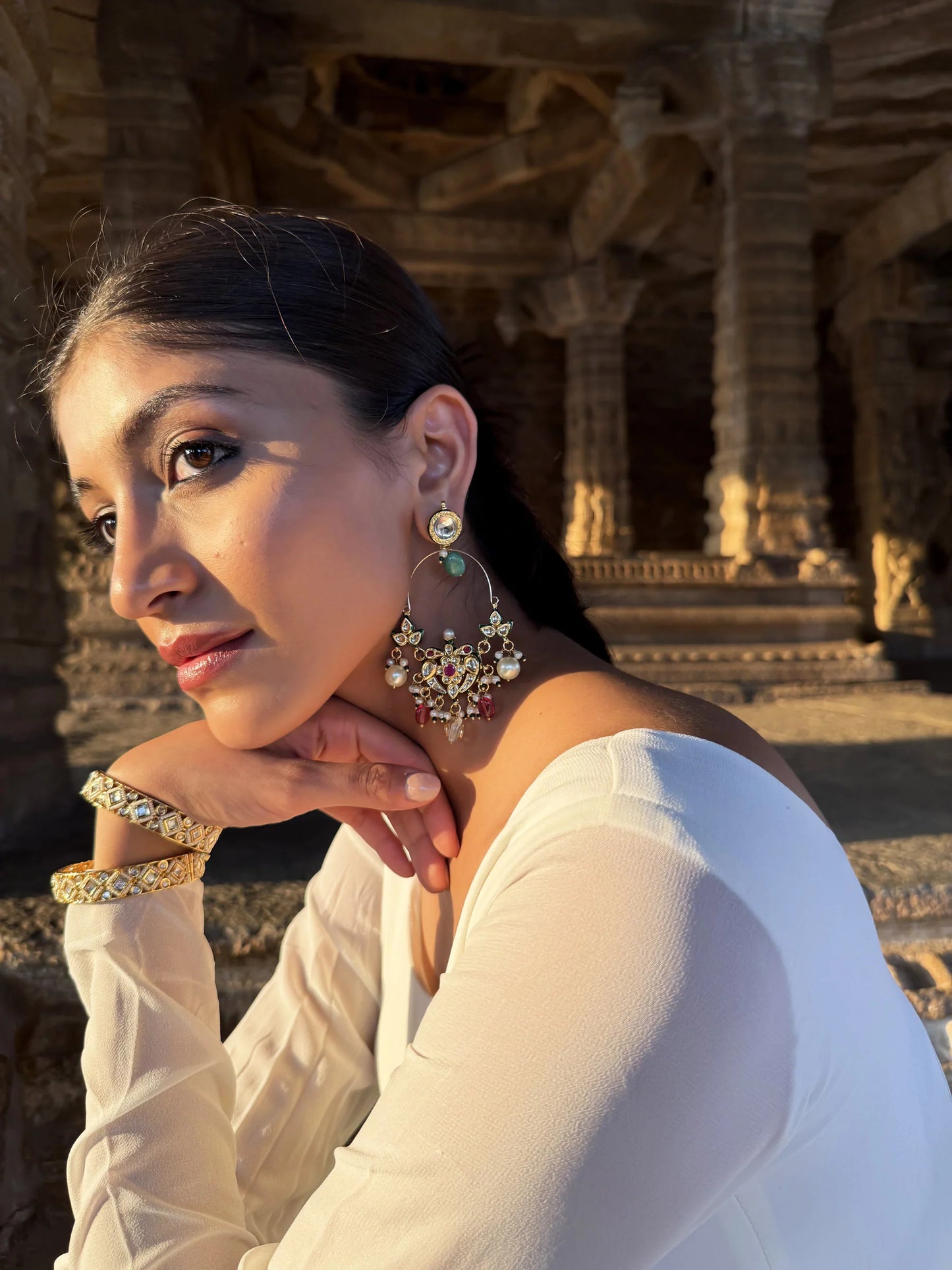 Elegant portrait of a woman in a white dress adorned with statement earrings, posed in a historic stone temple setting with warm sunlight

