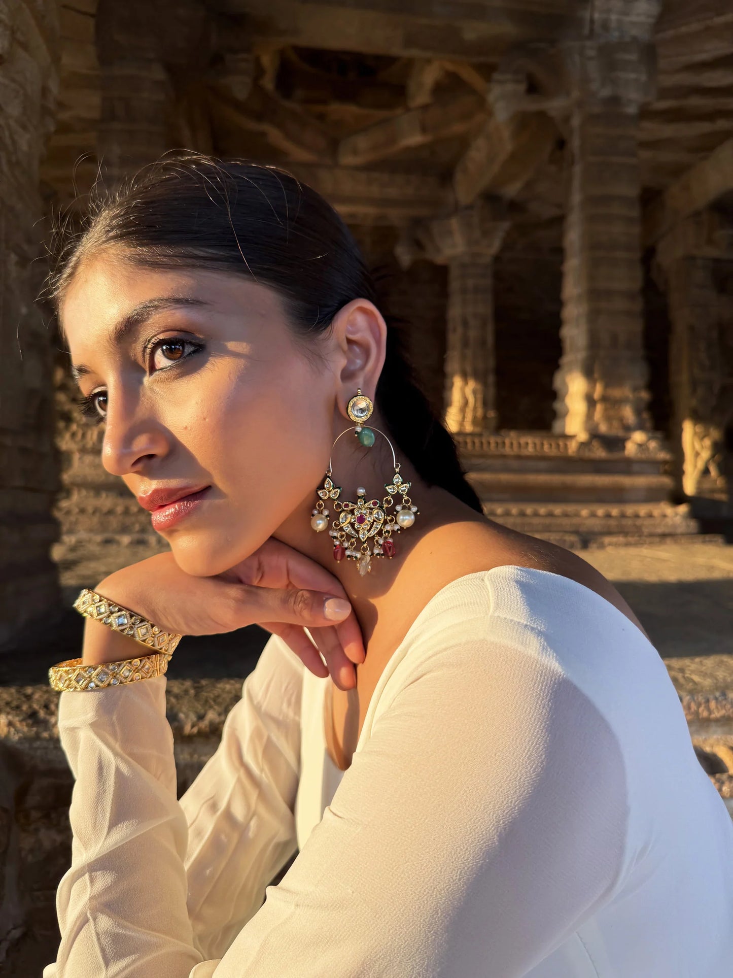 Cultural fashion portrait of a woman wearing detailed earrings and bangles, set against heritage stone architecture in warm light

