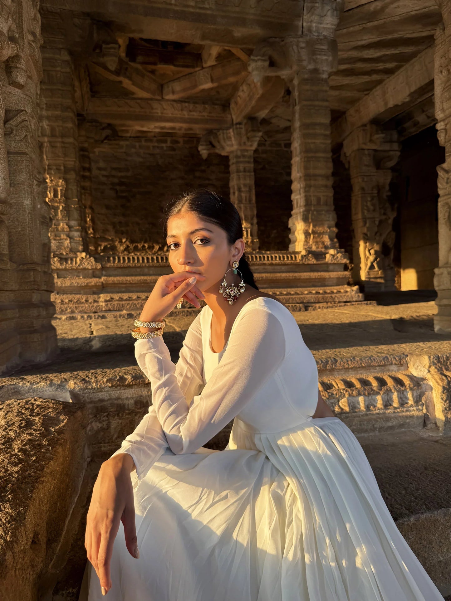 Golden hour portrait of a woman seated on stone steps, showcasing intricate earrings and bracelet in a heritage setting
