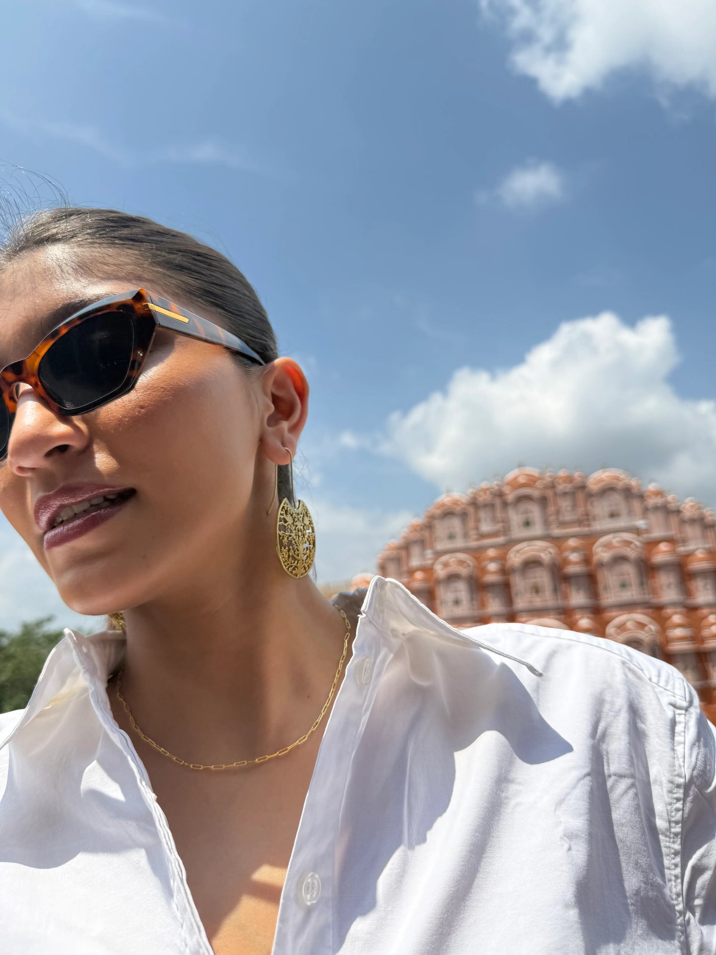 Travel fashion shot of woman wearing sunglasses and gold earrings near historic monument
