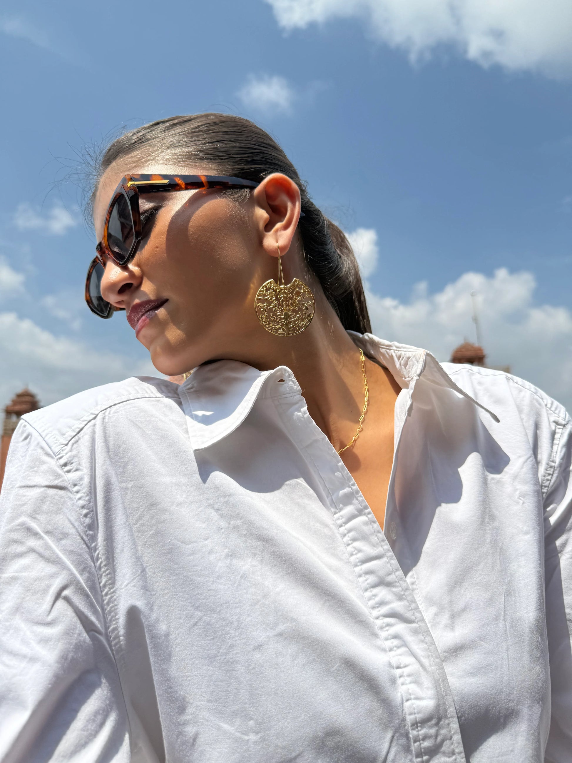 Luxury fashion image featuring gold earrings and sunglasses with heritage architecture setting
