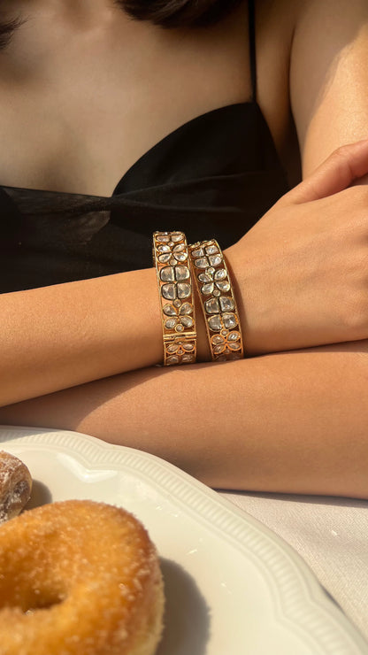 Close-up of a wrist wearing a gold bracelet with floral clusters of clear stones set in a sleek, polished frame.
