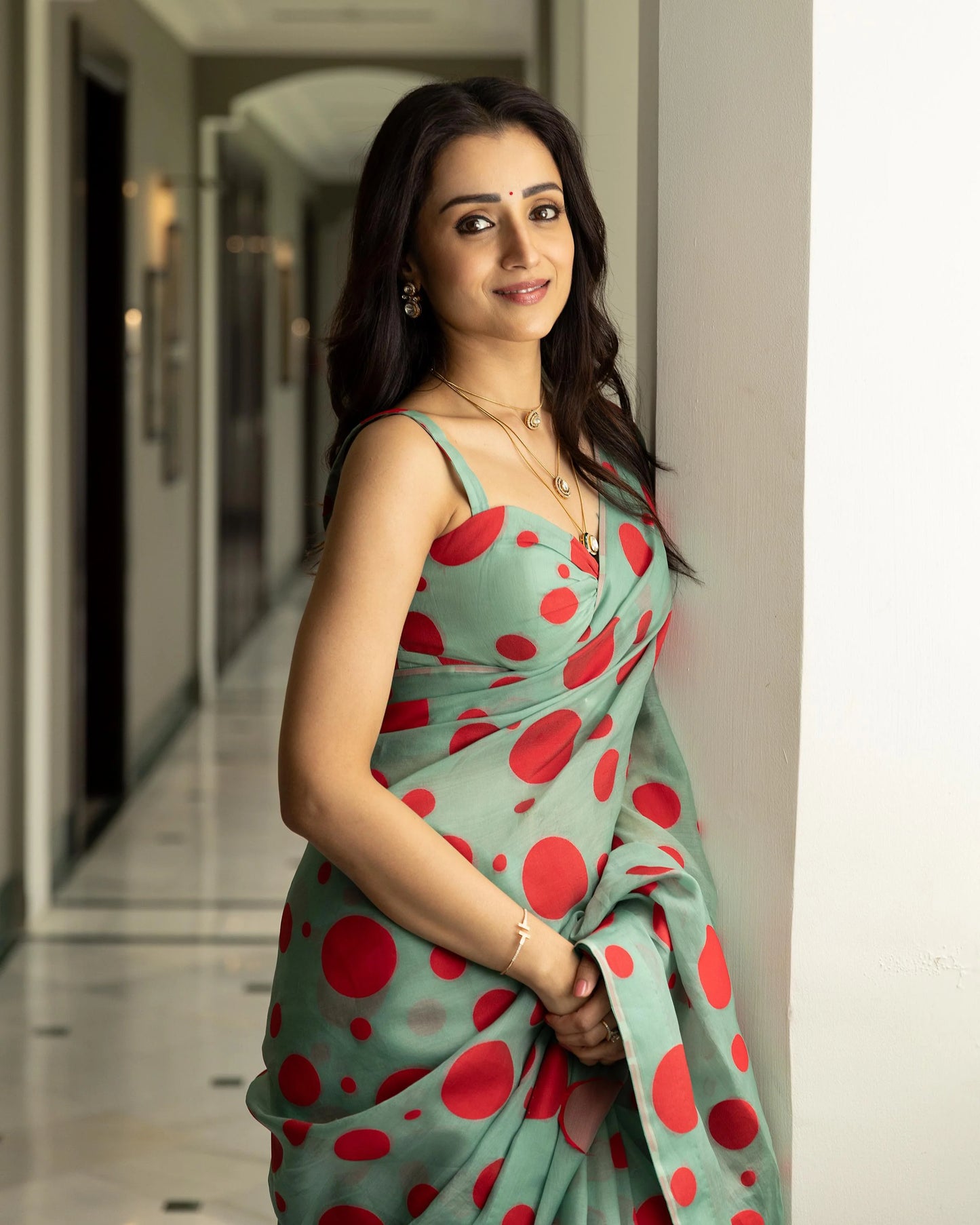 Woman in a green saree with red polka dots wearing layered gold necklaces and earrings, leaning against a wall
