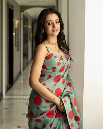 Woman in a green saree with red polka dots wearing layered gold necklaces and earrings, leaning against a wall
