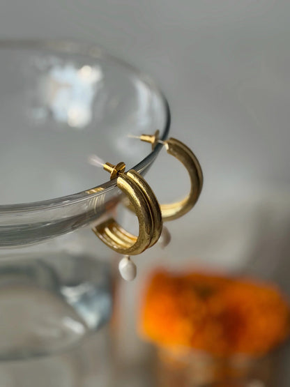 Elegant gold hoop earrings with minimal pearl accent displayed on glass with blurred background
