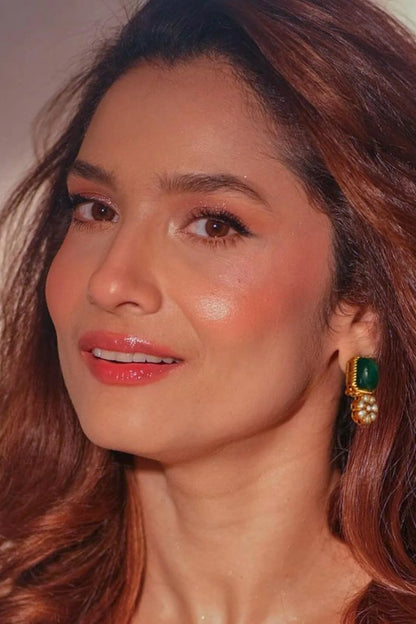 Close-up of a woman’s ear showcasing a gold earring with a deep green gemstone and pearl cluster detail in a warm-toned setting.
