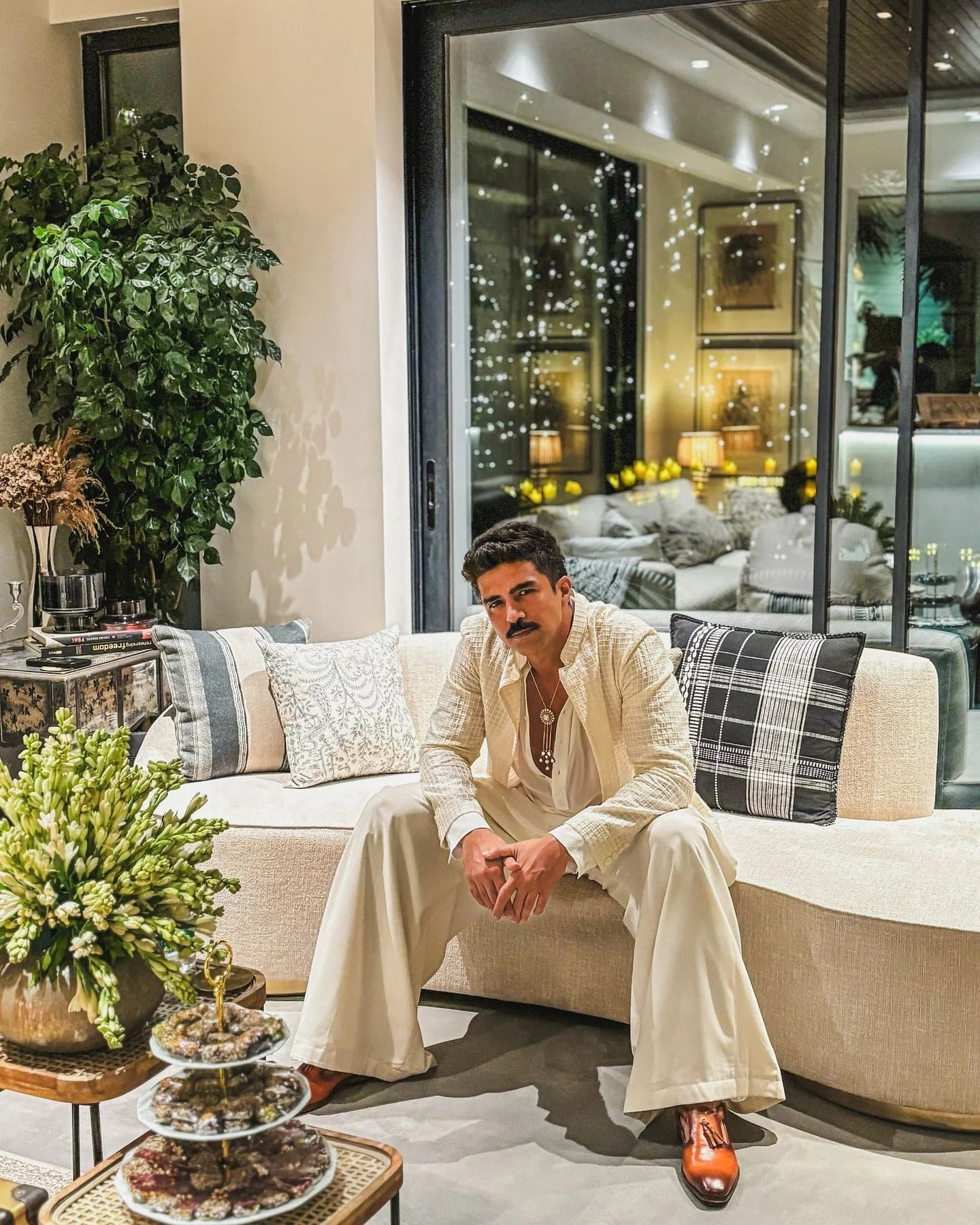 Man sitting on a modern beige sofa in a luxury living room with warm lighting and indoor plants at night
