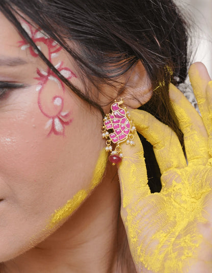 Detailed shot of a decorative earring with bright pink stone segments, green accents, and dangling pearls, worn against a vibrant, artistic backdrop.
