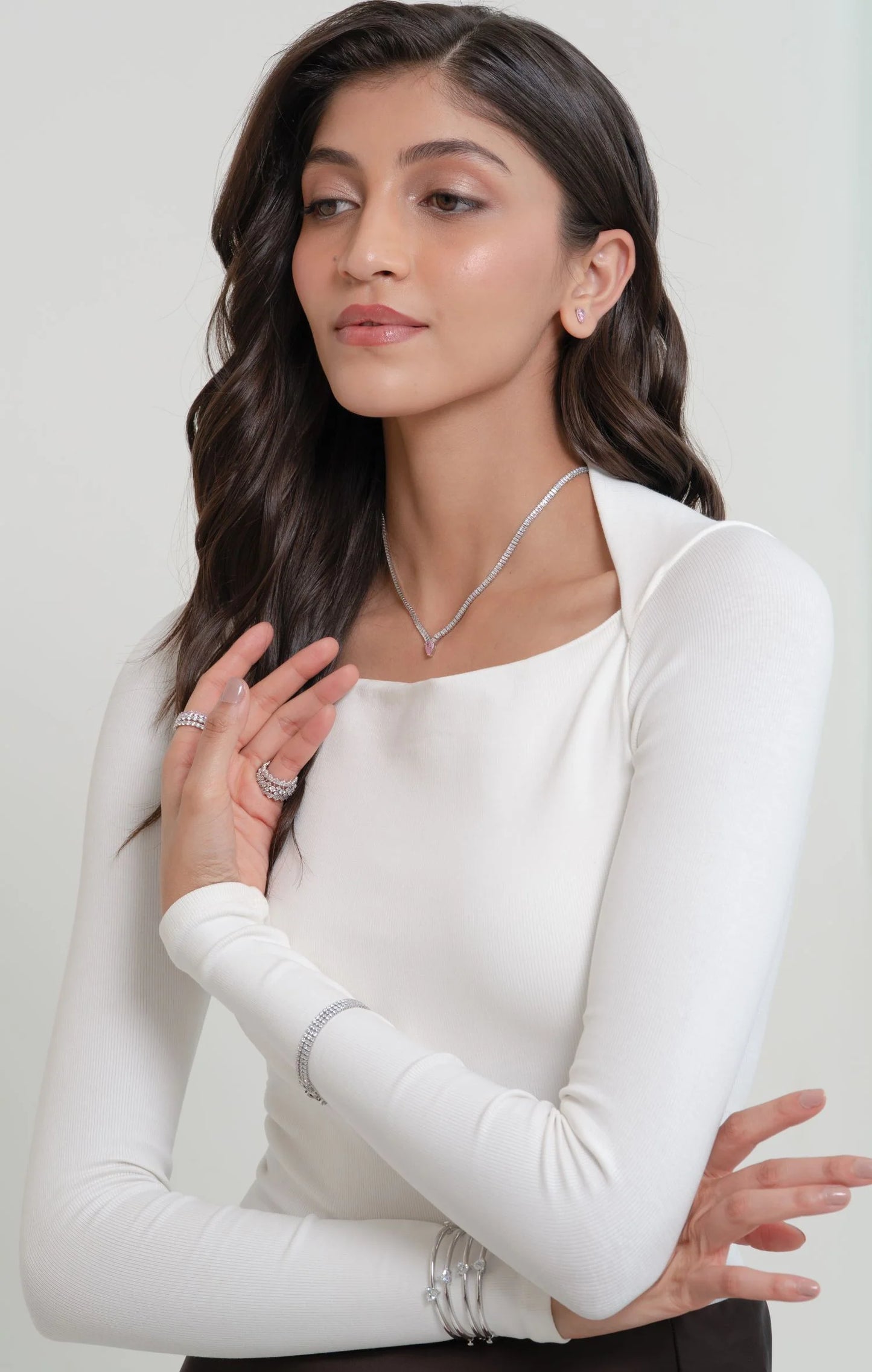 Fashion portrait of a woman wearing layered diamond rings, a slim bracelet, and a sparkling pendant necklace against a clean background.
