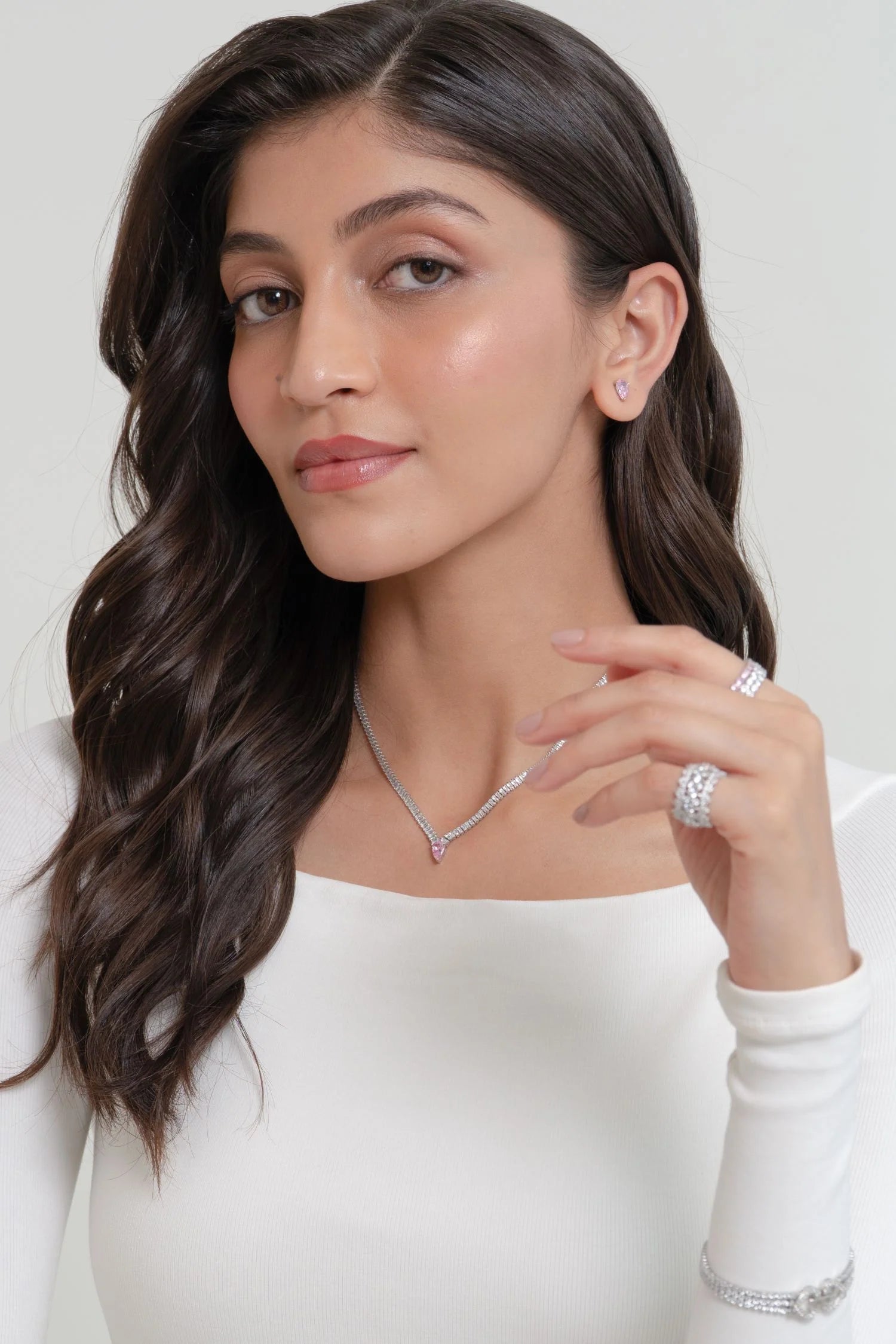 Portrait of a woman wearing a white top, styled with a diamond pendant necklace, stud earrings, rings, and a delicate bracelet.
